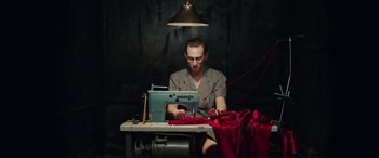 Movie still from “In Fabric” (2018), directed by Peter Strickland – A man sitting in front of a sewing machine on top of a table; Medium shot, High angle
