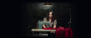 Movie still from “In Fabric” (2018), directed by Peter Strickland – A woman sitting at a sewing machine with a red cloth; Medium shot, High angle