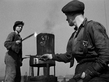 Movie still from “In Which We Serve” (1942), directed by Noël Coward – Two men are standing in front of a chair; Medium shot, Low angle