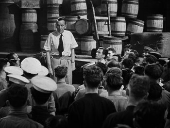Movie still from “In Which We Serve” (1942), directed by Noël Coward – An old photo of a group of people gathered in a room; Wide shot, High angle