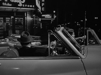 Movie still from “In a Lonely Place” (1950), directed by Nicholas Ray – A man in a hat driving a car in a city; Wide shot, Over the shoulder angle