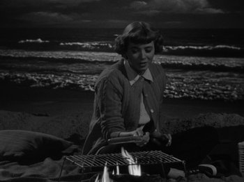 Movie still from “In a Lonely Place” (1950), directed by Nicholas Ray – A black and white photo of a woman cooking on the beach; Medium shot, High angle