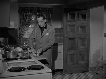 Movie still from “In a Lonely Place” (1950), directed by Nicholas Ray – A black and white photo of a man standing in a kitchen preparing a meal; Medium shot, Low angle