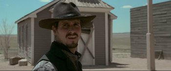 Movie still from “In a Valley of Violence” (2016), directed by Ti West – A man wearing a hat standing in front of a building; Close Up shot, Over the shoulder angle