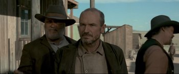 Movie still from “In a Valley of Violence” (2016), directed by Ti West – A man with a beard is standing next to another man; Medium shot, Low angle