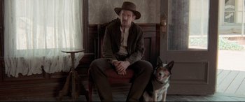 Movie still from “In a Valley of Violence” (2016), directed by Ti West – A man sitting on a chair next to a dog; Medium shot, Low angle