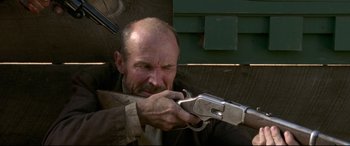 Movie still from “In a Valley of Violence” (2016), directed by Ti West – A man holding a gun while wearing a jacket; Close Up shot, Low angle