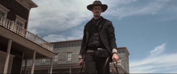 Movie still from “In a Valley of Violence” (2016), directed by Ti West – A man in a hat holding a whip in front of a building; Medium shot, Low angle