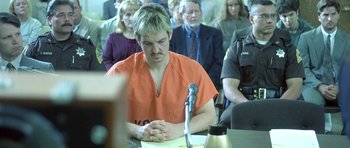 Movie still from “In the Bedroom” (2001), directed by Todd Field – A man in an orange shirt sitting in front of a microphone in a courtroom; Medium shot, Over the shoulder angle