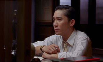 Movie still from “In the Mood for Love” (2000), directed by Kar-Wai Wong – A man sitting at a table with his hands folded on his chest; Close Up shot, Over the shoulder angle