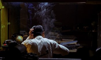 Movie still from “In the Mood for Love” (2000), directed by Kar-Wai Wong – A man sitting in front of a table smoking a cigarette; Medium shot, High angle