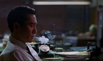 Movie still from “In the Mood for Love” (2000), directed by Kar-Wai Wong – A man sitting at a desk smoking a cigarette; Close Up shot, Low angle