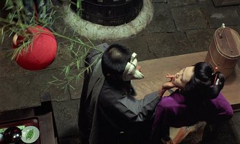 Movie still from “In the Realm of the Senses” (1976), directed by Nagisa Ôshima – A man with a mask is sitting on a bench with a woman; Medium shot, High angle