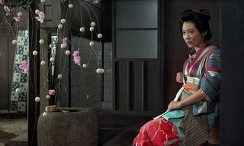 Movie still from “In the Realm of the Senses” (1976), directed by Nagisa Ôshima – A woman sitting on a chair in front of a plant; Medium shot, Low angle