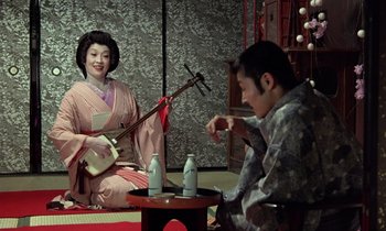 Movie still from “In the Realm of the Senses” (1976), directed by Nagisa Ôshima – A woman sitting next to a man holding a guitar; Medium shot, Over the shoulder angle