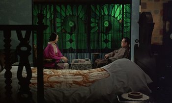 Movie still from “In the Realm of the Senses” (1976), directed by Nagisa Ôshima – A man and a woman sitting on a bed in front of a stained glass window; Wide shot, High angle