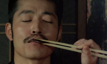 Movie still from “In the Realm of the Senses” (1976), directed by Nagisa Ôshima – A man holding a pair of chopsticks in his mouth; Extreme Close Up shot, Low angle