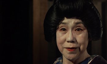Movie still from “In the Realm of the Senses” (1976), directed by Nagisa Ôshima – An older woman wearing a black wig and a blue shirt; Close Up shot, Over the shoulder angle