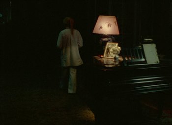 Movie still from “India Song” (1975), directed by Marguerite Duras – A woman standing in front of a table with a lamp on top of it; Wide shot, Over the shoulder angle