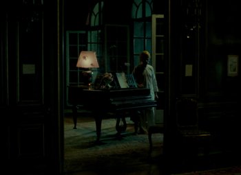 Movie still from “India Song” (1975), directed by Marguerite Duras – A man standing in front of a grand piano in a dark room; Wide shot, High angle