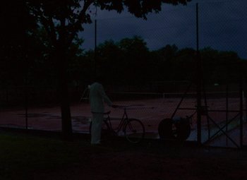 Movie still from “India Song” (1975), directed by Marguerite Duras – A person standing next to a bike in a field; Extreme Wide shot, High angle