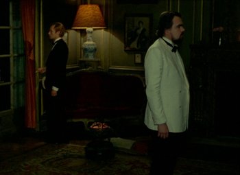 Movie still from “India Song” (1975), directed by Marguerite Duras – Two men dressed in suits stand in front of a fireplace; Wide shot, Over the shoulder angle