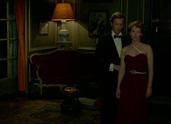 Movie still from “India Song” (1975), directed by Marguerite Duras – A man and a woman standing in front of a couch in a living room; Wide shot, High angle