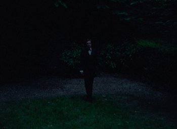 Movie still from “India Song” (1975), directed by Marguerite Duras – A man in a suit and tie standing in the dark; Wide shot, High angle