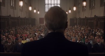 Movie still from “Indignation” (2016), directed by James Schamus – An old man is standing in front of a crowd of people; Medium shot, Low angle