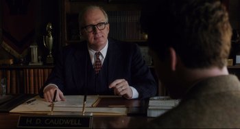 Movie still from “Indignation” (2016), directed by James Schamus – An older man sitting at a desk in a suit and tie; Medium shot, Over the shoulder angle