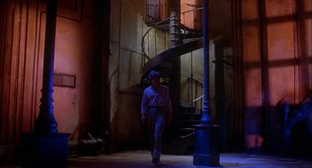 Movie still from “Inferno” (1980), directed by Dario Argento – A man walking in a dimly - lit room next to a spiral staircase; Wide shot, Low angle