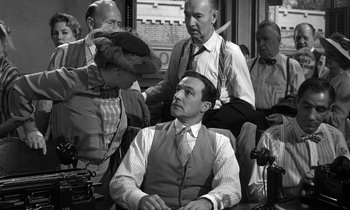Movie still from “Inherit the Wind” (1960), directed by Stanley Kramer – A man sitting in a chair in front of a group of people; Medium shot, Over the shoulder angle