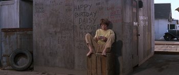 Movie still from “Inside Daisy Clover” (1965), directed by Robert Mulligan – A person sitting on a wooden box in front of a wall; Wide shot, High angle