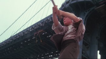Movie still from “Insignificance” (1985), directed by Nicolas Roeg – A young boy swinging a baseball bat in front of a bridge; Medium shot, Low angle