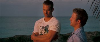 Movie still from “Into the Blue” (2005), directed by John Stockwell – A man standing next to a body of water with his arms crossed; Medium shot, Over the shoulder angle