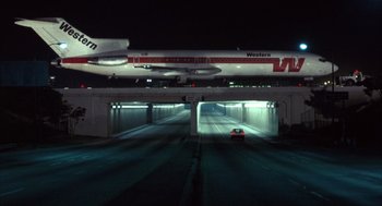 Movie still from “Into the Night” (1985), directed by John Landis – An airplane is flying over an underpass at night; Extreme Wide shot, Low angle