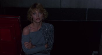 Movie still from “Into the Night” (1985), directed by John Landis – A woman with her arms crossed in a dark room; Medium shot, Low angle