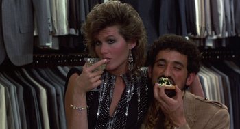 Movie still from “Into the Night” (1985), directed by John Landis – A man and a woman are eating a doughnut together; Medium shot, Low angle