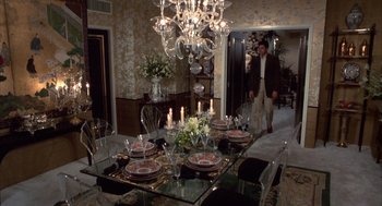 Movie still from “Into the Night” (1985), directed by John Landis – A dining room table set for a meal with a man standing in the background; Wide shot, High angle