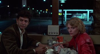 Movie still from “Into the Night” (1985), directed by John Landis – A man and a woman sitting at a table in front of a window; Medium shot, Low angle