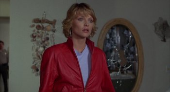 Movie still from “Into the Night” (1985), directed by John Landis – A woman wearing a red jacket standing in a room; Medium shot, Over the shoulder angle