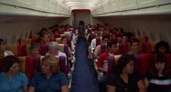Movie still from “Into the Night” (1985), directed by John Landis – A group of people sitting on a plane in the middle of the aisle; Wide shot, High angle