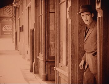Movie still from “Intolerance” (1916), directed by D.W. Griffith – A man in a suit and hat leaning against a building; Wide shot, High angle