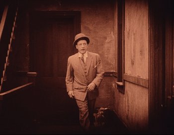 Movie still from “Intolerance” (1916), directed by D.W. Griffith – A man in a suit and hat standing next to a door; Wide shot, High angle