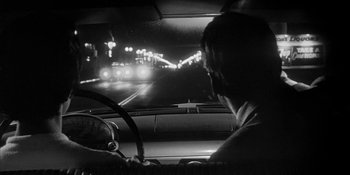 Movie still from “Invasion of the Body Snatchers” (1956), directed by Don Siegel – A man driving a car at night on the street; Medium shot, Over the shoulder angle