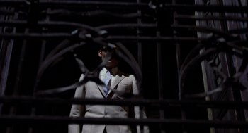 Movie still from “Investigation of a Citizen Above Suspicion” (1970), directed by Elio Petri – A man wearing a suit and tie standing in front of a metal fence; Medium shot, Low angle