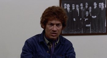 Movie still from “Investigation of a Citizen Above Suspicion” (1970), directed by Elio Petri – A man in a blue jacket sitting in front of a picture; Close Up shot, Low angle