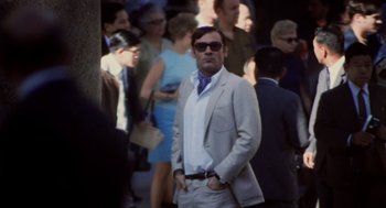 Movie still from “Investigation of a Citizen Above Suspicion” (1970), directed by Elio Petri – A man standing in a crowd of people wearing a suit; Medium shot, Low angle