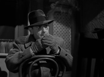 Movie still from “Invisible Agent” (1942), directed by Edwin L. Marin – A man in a hat smoking a cigarette in a chair; Medium shot, Over the shoulder angle