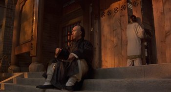 Movie still from “Iron Monkey” (1993), directed by Woo-Ping Yuen – A man sitting on the steps in front of a wooden building; Wide shot, Low angle